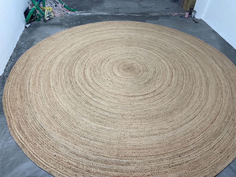 K&ouml;nnte beinhalten: Ein gro&szlig;er, runder Jute-Teppich mit einem konzentrischen Spiralmuster. Der Teppich hat eine nat&uuml;rliche, beige Farbe und scheint aus gewebten Fasern zu bestehen. Der Teppich liegt auf einem Betonboden.
