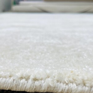 May include: Close-up of a white, textured rug. The rug has a soft, plush appearance with a visible border. The fibers create a subtle pattern, suggesting a comfortable and inviting texture. The rug's neutral color makes it versatile for various interior designs.