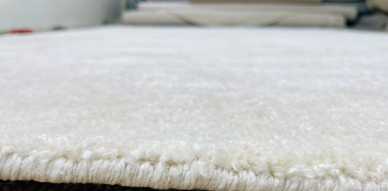 May include: Close-up of a white, textured rug. The rug has a soft, plush appearance with a visible border. The fibers create a subtle pattern, suggesting a comfortable and inviting texture. The rug's neutral color makes it versatile for various interior designs.