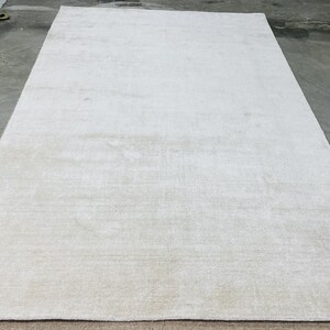 May include: A large, rectangular, off-white rug with a subtle, textured pattern. The rug is laid out on a concrete floor, showcasing its size and neutral color palette. The rug's edges are neatly finished, suggesting a high-quality construction.