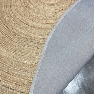 K&ouml;nnte beinhalten: Ein runder Teppich aus Naturfasern mit einem Spiralmuster. Die hellbraune, gewebte Oberfl&auml;che des Teppichs kontrastiert mit der grauen R&uuml;ckseite. Das Design und die Textur des Teppichs schaffen eine warme und einladende &Auml;sthetik.