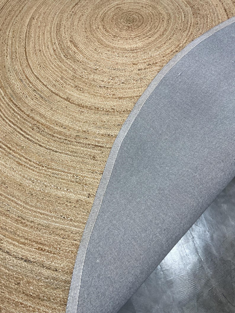 K&ouml;nnte beinhalten: Ein runder Teppich aus Naturfasern mit einem Spiralmuster. Die hellbraune, gewebte Oberfl&auml;che des Teppichs kontrastiert mit der grauen R&uuml;ckseite. Das Design und die Textur des Teppichs schaffen eine warme und einladende &Auml;sthetik.
