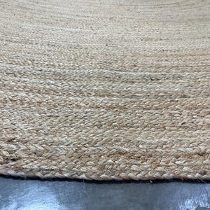 K&ouml;nnte beinhalten: Nahaufnahme eines runden Teppichs aus nat&uuml;rlichem Jute. Der Teppich hat eine gewebte Textur mit einem geflochtenen Rand. Die Farbe ist ein helles Beige mit Farbvariationen, wodurch ein strukturiertes Aussehen entsteht. Der Teppich liegt auf einer grauen Oberfl&auml;che.