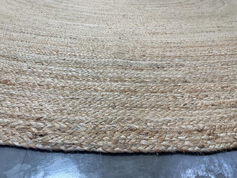K&ouml;nnte beinhalten: Nahaufnahme eines runden Teppichs aus nat&uuml;rlichem Jute. Der Teppich hat eine gewebte Textur mit einem geflochtenen Rand. Die Farbe ist ein helles Beige mit Farbvariationen, wodurch ein strukturiertes Aussehen entsteht. Der Teppich liegt auf einer grauen Oberfl&auml;che.
