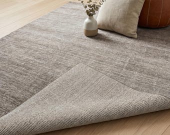 May include: A rectangular area rug with a textured, heathered gray surface and a folded corner revealing a lighter, woven underside. The rug is placed on a light wood floor alongside a beige throw pillow and a small white vase.