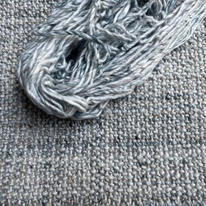 May include: A close-up shot of a skein of light blue and white yarn resting on a textured gray fabric. The yarn is loosely wound, showing the individual strands. The fabric has a woven appearance, with a speckled pattern.