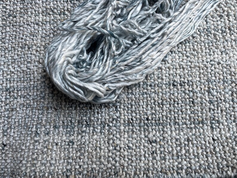 May include: A close-up shot of a skein of light blue and white yarn resting on a textured gray fabric. The yarn is loosely wound, showing the individual strands. The fabric has a woven appearance, with a speckled pattern.