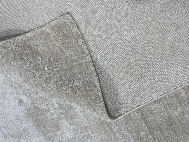 May include: Close-up of a light beige rug with a textured surface. The rug is folded over, revealing the underside and the edge of the rug. The rug appears to be made of a soft material.