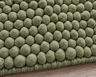 May include: A sage green rug featuring a dense, textured surface composed of numerous small, rounded, felted spheres arranged in a repeating pattern, finished with a thick, matching green braided border along the bottom edge.