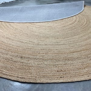 K&ouml;nnte beinhalten: Ein runder Jute-Teppich in Naturbeige mit hellgrauer R&uuml;ckseite. Die Oberfl&auml;che des Teppichs hat ein strukturiertes, gewebtes Aussehen mit konzentrischen kreisf&ouml;rmigen Mustern. Die R&uuml;ckseite ist oben im Bild sichtbar.