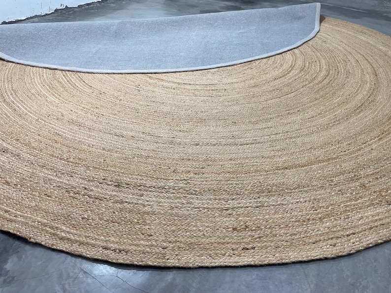 K&ouml;nnte beinhalten: Ein runder Jute-Teppich in Naturbeige mit hellgrauer R&uuml;ckseite. Die Oberfl&auml;che des Teppichs hat ein strukturiertes, gewebtes Aussehen mit konzentrischen kreisf&ouml;rmigen Mustern. Die R&uuml;ckseite ist oben im Bild sichtbar.