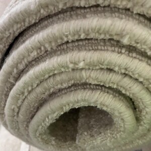 May include: Close-up of a rolled-up, light beige rug. The rug has a textured, looped pile, creating a soft, plush appearance. The edges of the rug are visible, showing the layers of the rolled material. The neutral color suggests it would complement various interior design styles.