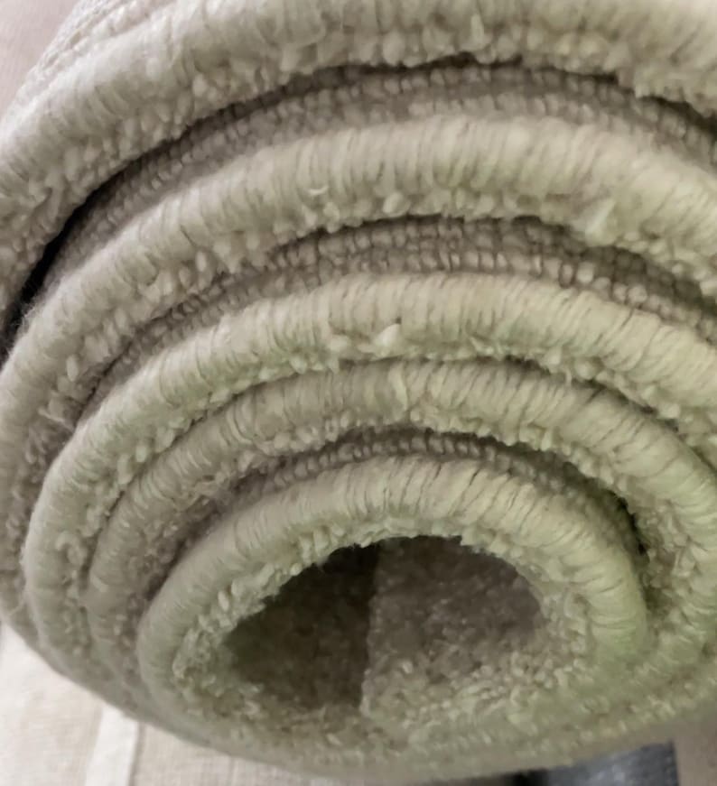 May include: Close-up of a rolled-up, light beige rug. The rug has a textured, looped pile, creating a soft, plush appearance. The edges of the rug are visible, showing the layers of the rolled material. The neutral color suggests it would complement various interior design styles.