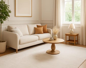 May include: A cream-colored sofa with two orange and one white throw pillow, a round wooden coffee table with a vase, a small wooden side table with a lamp, a potted plant, and a large cream area rug on a light wood herringbone floor.
