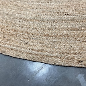 K&ouml;nnte beinhalten: Nahaufnahme eines runden, nat&uuml;rlichen Jute-Teppichs. Der Teppich hat eine gewebte Textur mit hellbrauner Farbe. Der Rand des Teppichs ist sichtbar und zeigt das geflochtene Detail. Der Teppich liegt auf einer dunkelgrauen Oberfl&auml;che.
