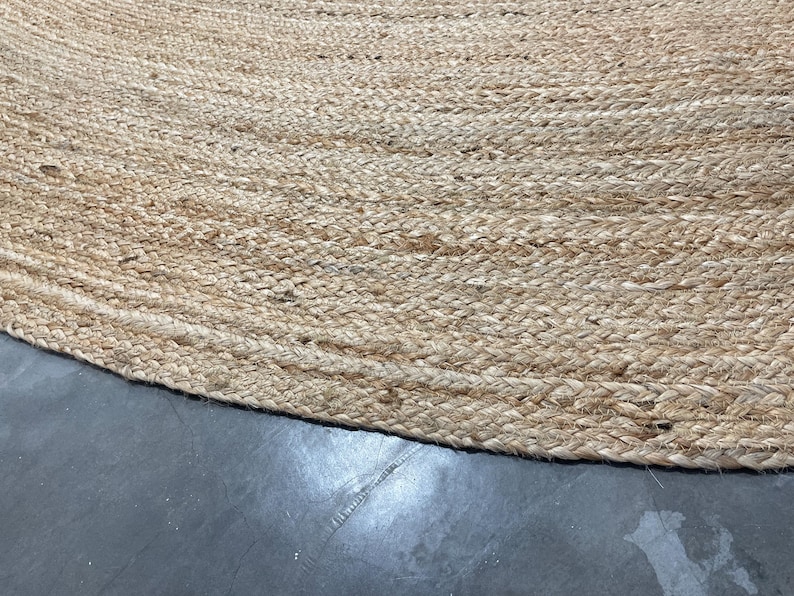 K&ouml;nnte beinhalten: Nahaufnahme eines runden, nat&uuml;rlichen Jute-Teppichs. Der Teppich hat eine gewebte Textur mit hellbrauner Farbe. Der Rand des Teppichs ist sichtbar und zeigt das geflochtene Detail. Der Teppich liegt auf einer dunkelgrauen Oberfl&auml;che.