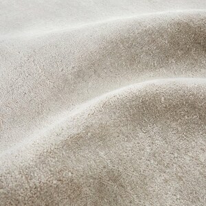 May include: Close-up of a textured, light beige fabric with a soft, plush appearance. The fabric has a slightly wavy pattern, creating shadows and highlights across its surface. The material appears to be thick and possibly used for upholstery or textiles.