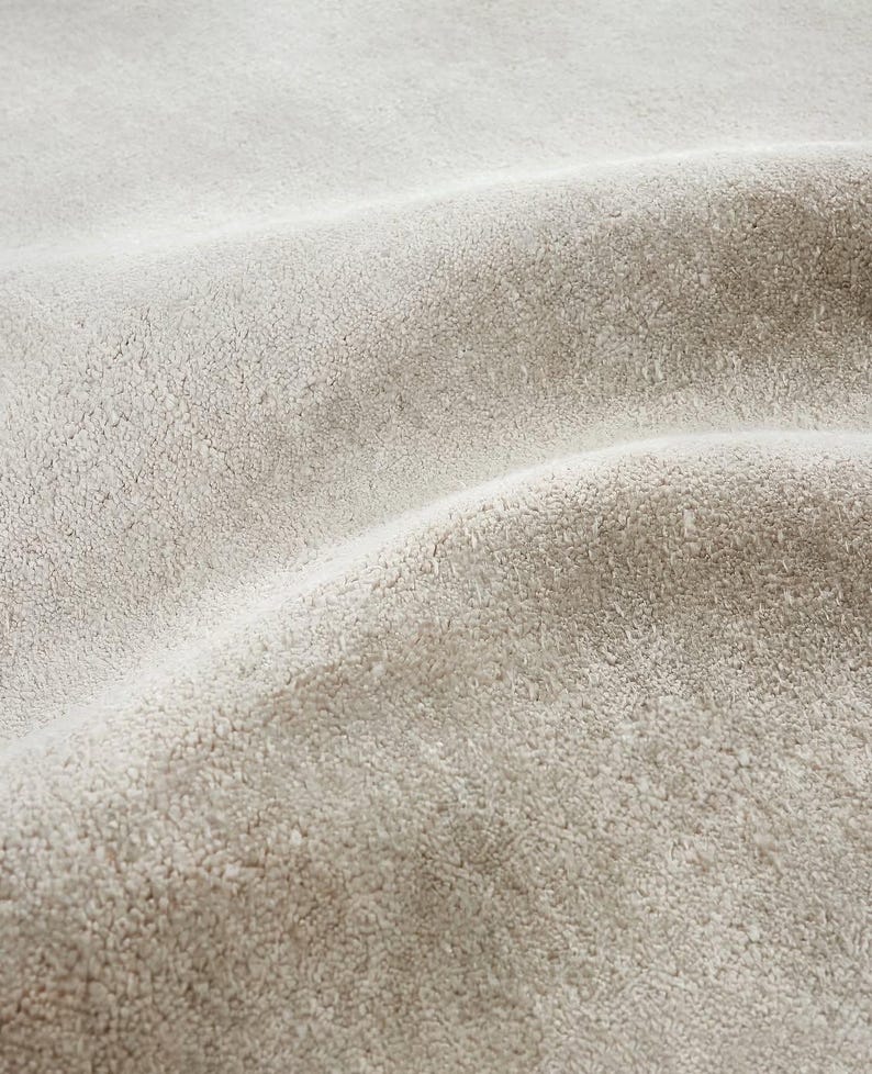 May include: Close-up of a textured, light beige fabric with a soft, plush appearance. The fabric has a slightly wavy pattern, creating shadows and highlights across its surface. The material appears to be thick and possibly used for upholstery or textiles.