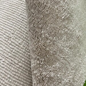 May include: Close-up of two sections of a light beige rug. One section has a ribbed pattern, while the other has a plush, textured surface. The rug appears soft and inviting, suitable for home decor.