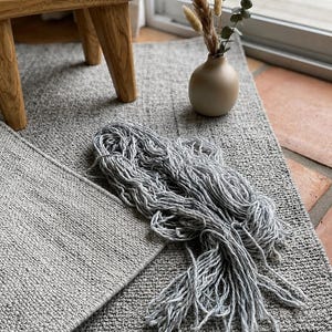 May include: A neutral-toned interior scene featuring a small wooden stool, a beige vase with dried plants, and a gray rug. A bundle of gray yarn rests on the rug, suggesting a craft or textile theme. The setting is illuminated by natural light.