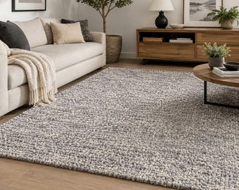 Puede incluir: Una alfombra rectangular que presenta una textura de tejido grueso con una paleta de colores variados en hilos gris carbón, gris claro y blanco roto. La alfombra tiene una construcción de pelo denso y rizado que crea una superficie uniforme.