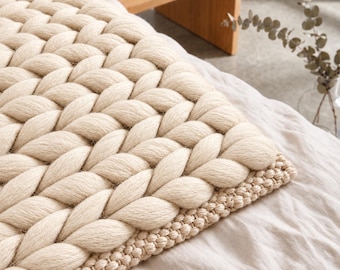 May include: A cream-colored chunky knit blanket featuring a large-scale braided pattern with a smaller, tight-knit border along the edge. The thick, soft yarn creates a textured surface that is draped over a white fabric base.