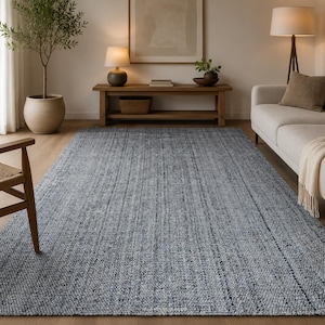 May include: A large, rectangular, woven area rug in shades of gray and blue. The rug has a textured appearance with vertical lines, adding visual interest. It is placed in a living room setting with a sofa, wooden furniture, and a potted plant.