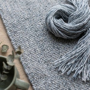 May include: A close-up of a light gray woven rug with a matching skein of yarn. The rug has a textured surface and a fringe of yarn. The yarn is a lighter shade of gray and is coiled on top of the rug. The scene is set on a wooden surface.