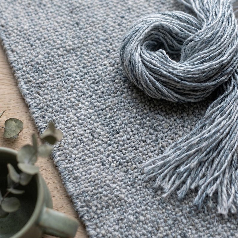 May include: A close-up of a light gray woven rug with a matching skein of yarn. The rug has a textured surface and a fringe of yarn. The yarn is a lighter shade of gray and is coiled on top of the rug. The scene is set on a wooden surface.
