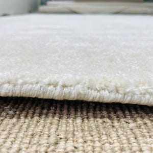 May include: A thick, off-white area rug with a soft, plush pile texture. The edge of the rug is finished with a tightly woven, matching off-white yarn binding that secures the fibers along the perimeter of the rectangular floor covering.