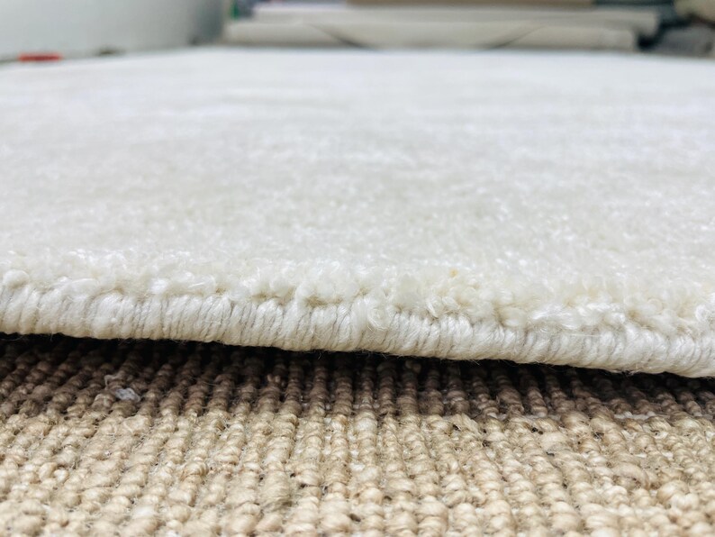May include: A thick, off-white area rug with a soft, plush pile texture. The edge of the rug is finished with a tightly woven, matching off-white yarn binding that secures the fibers along the perimeter of the rectangular floor covering.
