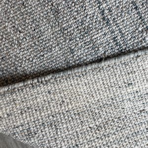 May include: Close-up of a gray and white woven rug. The rug has a textured surface with flecks of darker gray and blue. The edge of the rug is visible, showing the woven construction.
