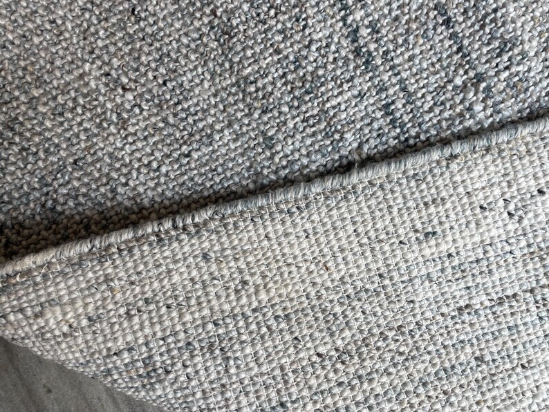 May include: Close-up of a gray and white woven rug. The rug has a textured surface with flecks of darker gray and blue. The edge of the rug is visible, showing the woven construction.