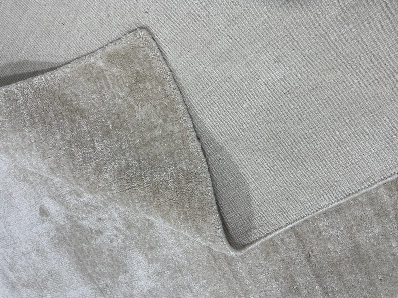 May include: Close-up of a light beige rug with a textured surface. The rug is folded over, revealing the underside and the edge of the rug. The rug appears to be made of a soft material.