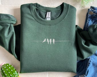 May include: A dark green sweatshirt with a crew neck and long sleeves. The sweatshirt features a minimalist design of four white birds perched on a thin white line. The brand tag is visible on the upper back.