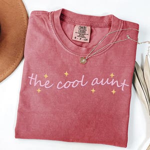 May include: A faded rose-coloured Comfort Colors t-shirt featuring the text "the cool aunt" in pink script, with yellow star accents. A silver necklace with a pendant is draped across the shirt.