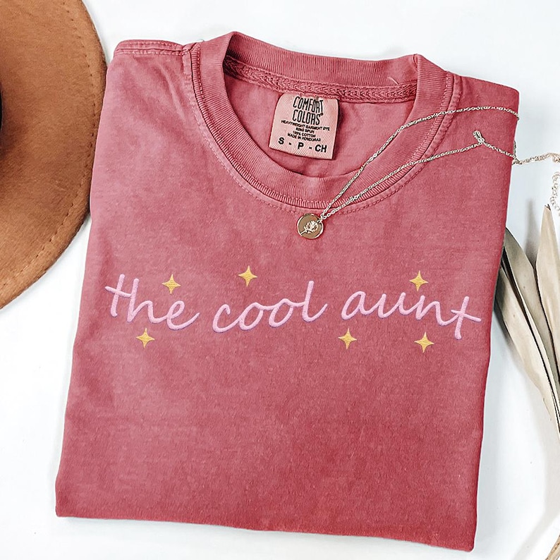 May include: A faded rose-coloured Comfort Colors t-shirt featuring the text "the cool aunt" in pink script, with yellow star accents. A silver necklace with a pendant is draped across the shirt.