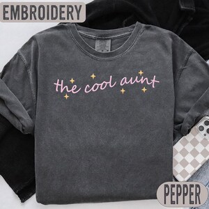 May include: A charcoal grey sweatshirt featuring the pink embroidered phrase "the cool aunt", with yellow star accents. The top reads "EMBROIDERY", and the bottom says "PEPPER". This casual sweatshirt is designed for comfort.