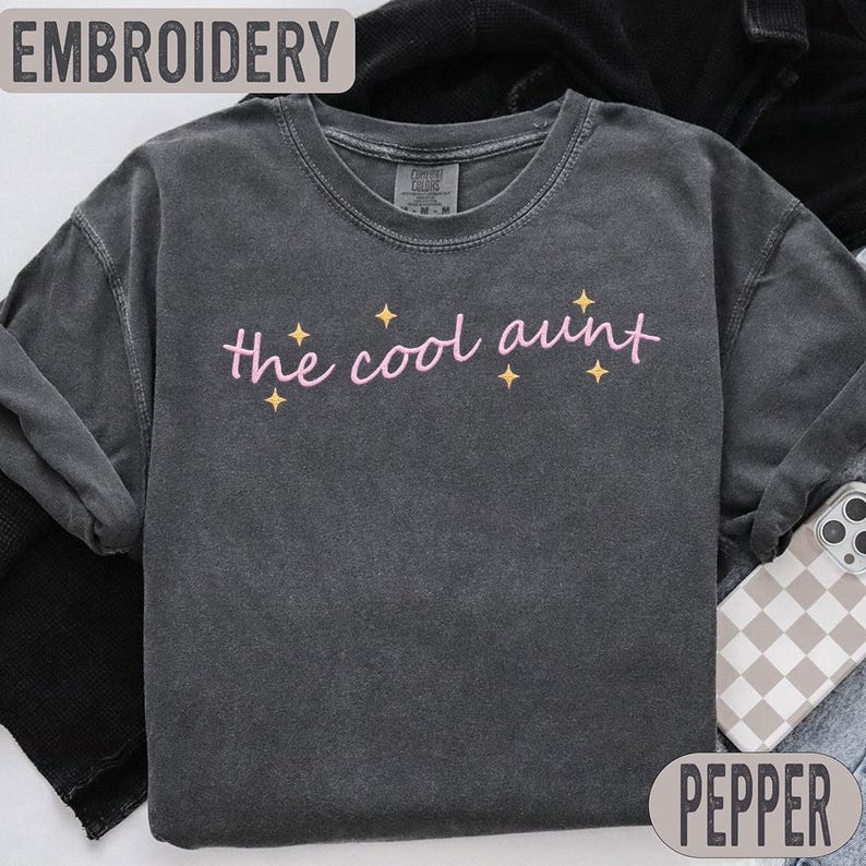 May include: A charcoal grey sweatshirt featuring the pink embroidered phrase "the cool aunt", with yellow star accents. The top reads "EMBROIDERY", and the bottom says "PEPPER". This casual sweatshirt is designed for comfort.