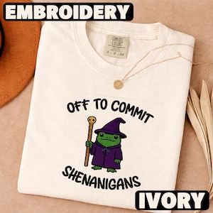 May include: Ivory-colored t-shirt with the word "EMBROIDERY" at the top. The shirt features a cartoon frog in a purple wizard outfit holding a staff, with the text "OFF TO COMMIT SHENANIGANS". The word "IVORY" is at the bottom.