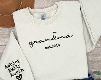 May include: Cream-colored sweatshirt with the word "grandma" in black script and "est. 2013" below it. The left sleeve has the names "Ashley, Kelly, Kevin" and a heart. The sweatshirt is folded.