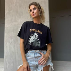 May include: A black short-sleeve t-shirt featuring a graphic of a ghost wearing a nurse cap and pushing a medical cart. The text "CODE BOO" is printed in gold above the ghost. The shirt is paired with light blue denim shorts with frayed hems.