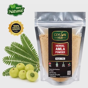 Op de afbeelding: Een zilveren stazak met 100g lichtbruin kruiden-amla-poeder, gelabeld Ceylon Hub. Het etiket bevat tekst, iconen en een QR-code. Naast de zak liggen vier groene amlavruchten en een takje met geveerde bladeren.