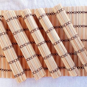 May include: A set of five light brown bamboo placemats, with four rolled into cylinders and one laid flat underneath. Each placemat features thin, vertical bamboo slats held together by dark brown thread in a repeating grid pattern.