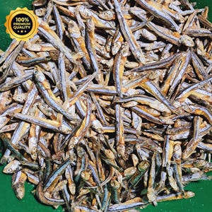 May include: A large pile of small, dried, silver and tan-coloured fish. A yellow circular emblem in the upper left corner features the text 100% PREMIUM QUALITY with three stars. The fish are arranged in a dense, overlapping cluster.