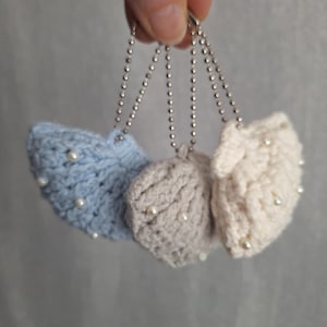 Puede incluir: Tres llaveros en forma de corazón tejidos a ganchillo en colores azul claro, gris y crema. Cada corazón está adornado con pequeñas perlas blancas y sujeto a una cadena de bolas plateada. Los llaveros son hechos a mano.