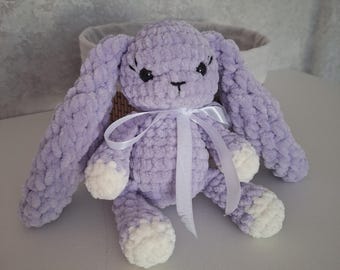 May include: A plush, light purple crocheted rabbit toy with long, floppy ears, black safety eyes, and white accents on the paws. The rabbit features a white satin ribbon tied in a bow around its neck and has a soft, textured yarn finish.