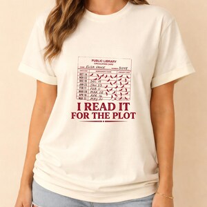 Op de afbeelding: Een crèmekleurig T-shirt met korte mouwen en een afbeelding van een vintage bibliotheekkaart. De kaart bevat de naam Eliza Vance, nummer 31048 en de tekst I READ IT FOR THE PLOT in donkerrode inkt met kleine rode markeringen.
