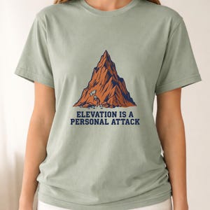 May include: A sage green short-sleeve t-shirt featuring a graphic of a large brown mountain with a small stick figure holding a flag at the base. Below the mountain, the text reads "ELEVATION IS A PERSONAL ATTACK" in dark blue capital letters.