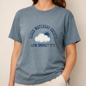Könnte beinhalten: Staubblaues T-Shirt mit Rundhalsausschnitt. Auf der Vorderseite befindet sich eine Grafik mit Wolke, Sonne und dem Text "CLOUD WATCHERS SOCIETY LOW IMPACT DIV." Das Shirt besteht aus weichem Material.
